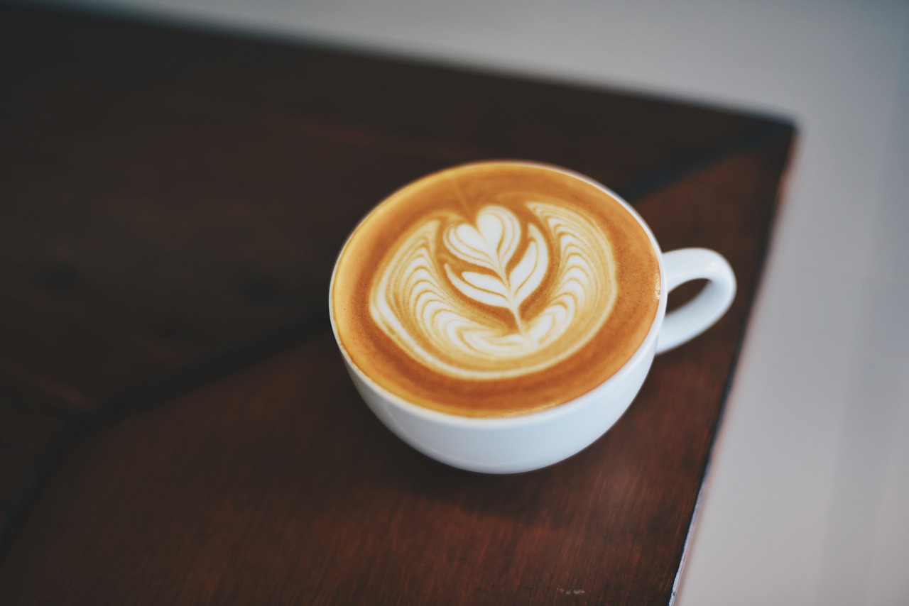 Kaffee steht in einer weißen Tasse auf einem dunkel Braunen Tisch. Der Milchschaum sieht aus wie eine Blume. 
